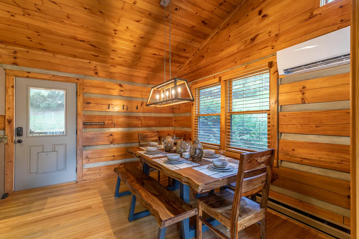 The scene centers on a rustic dining table beneath exposed wooden beams and a unique light fixture. Windows allow natural light to pour in, highlighting the inviting atmosphere and hinting at the natural surroundings just outside.