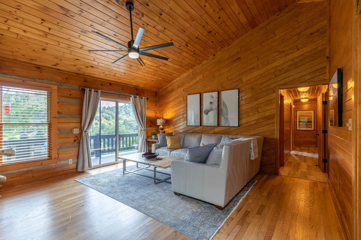 This image features a comfortable sofa and coffee table set in front of large windows with wooden walls and artwork. Through these expansive windows, the view outside hints at the surrounding natural scenery, bringing the outdoors in and inviting guests to relax while still feeling connected to the high country landscape.