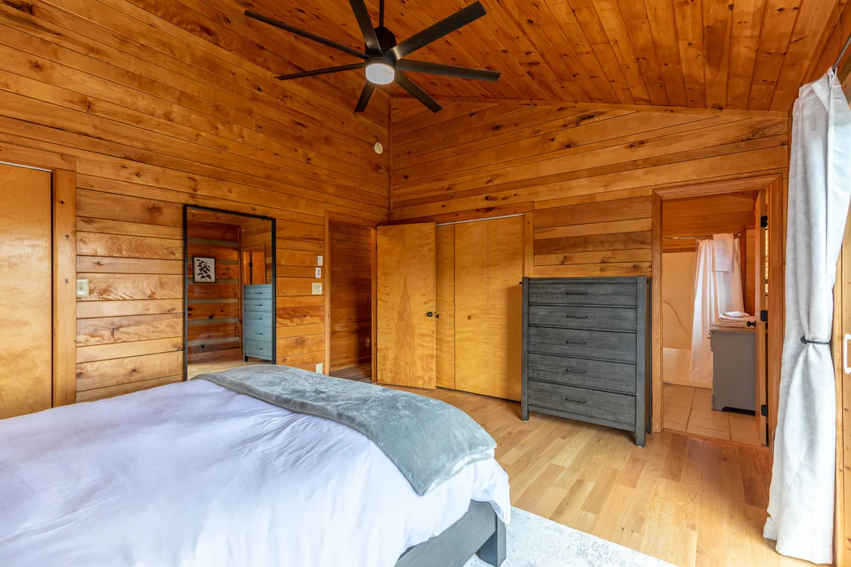 A tranquil bedroom with wooden walls, a cozy bed, and a large mirror, all bathed in natural light from a sizable window. The restful mood suggests a perfect place to unwind after days spent exploring the outdoors.