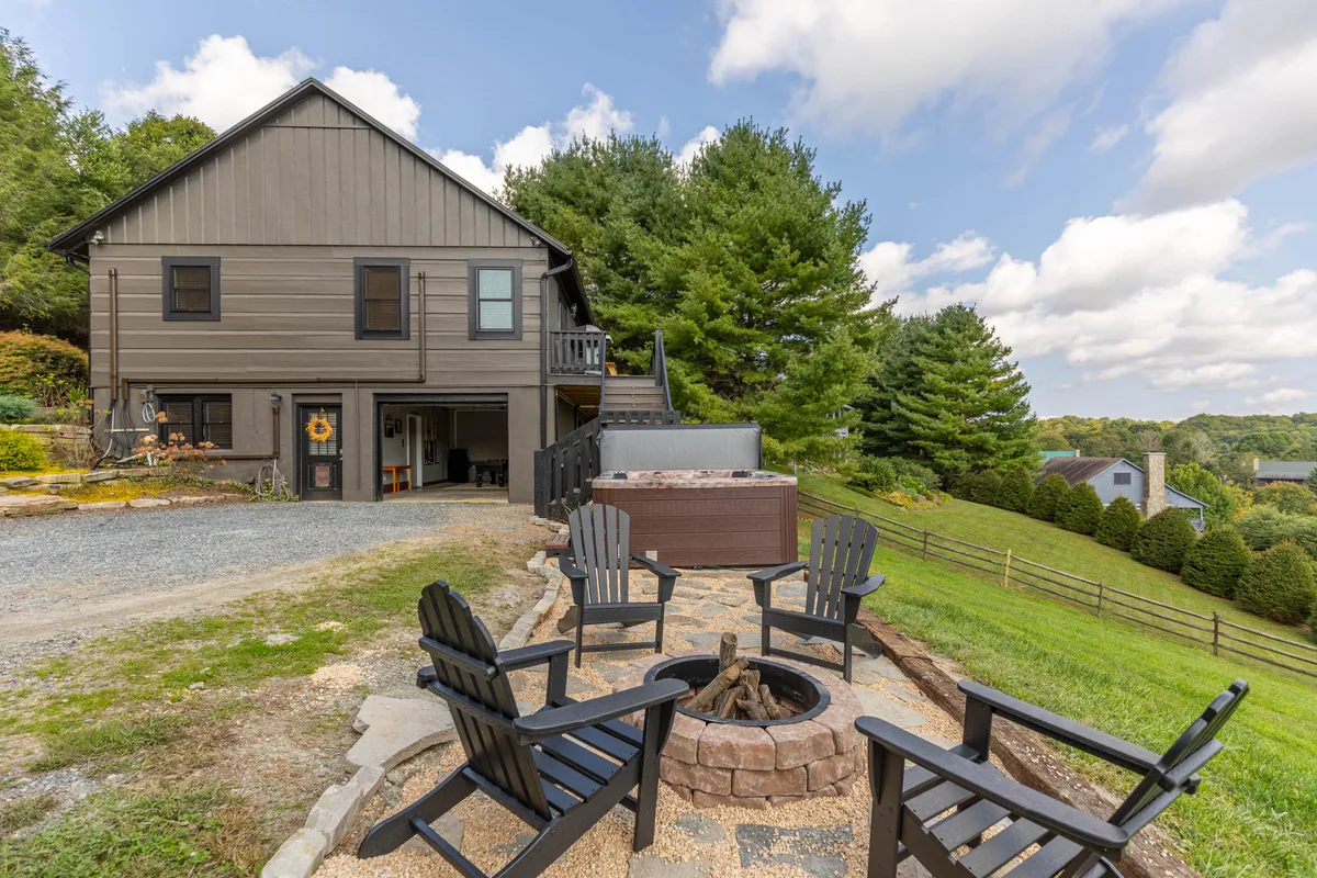 A wide view of the home's exterior featuring a cozy fire pit surrounded by Adirondack chairs, a spacious hot tub, and the house nestled amidst lush greenery. The scene captures outdoor gathering spots with plenty of seating, the hot tub, and a sense of welcoming communal warmth.