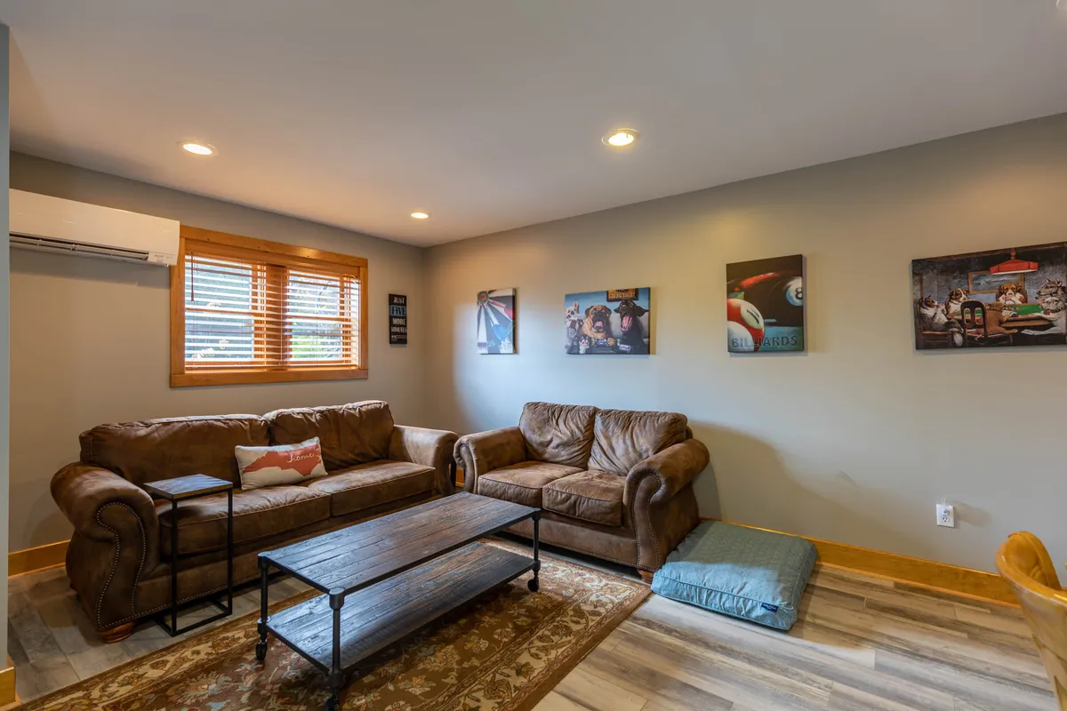 A cozy entertainment lounge featuring leather sofas, a coffee table, and wall art, with natural light streaming in from the window. The space is oriented for relaxation, movie nights, and socializing.