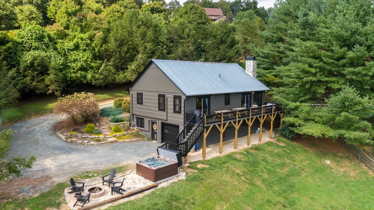 The image showcases the cabin's beautiful exterior nestled in the mountains, featuring a spacious deck, a steaming hot tub, an inviting fire pit area, and a lush, landscaped garden. The setting is peaceful, surrounded by natural beauty, with plenty of space for guests to gather and relax outdoors.