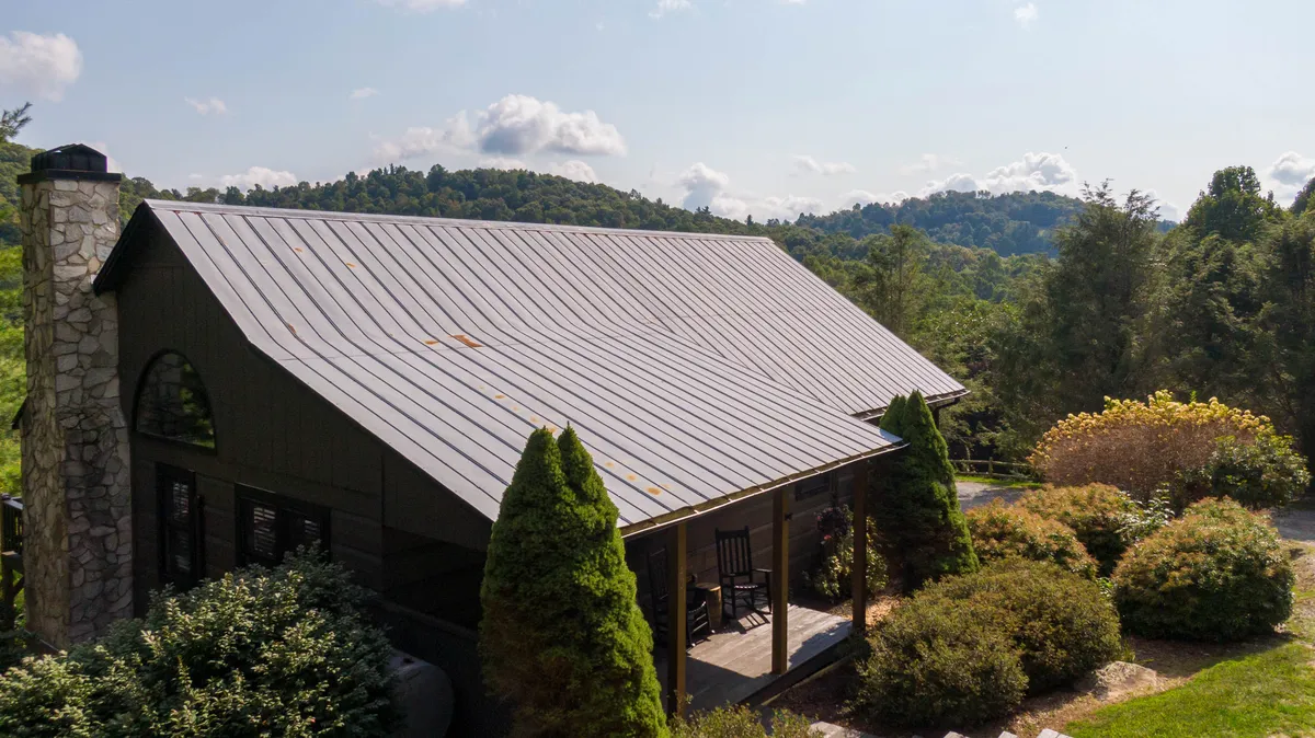 This image shows a black wooden house with a metal roof situated amidst lush greenery and overlooking mountain views. The porch offers a peaceful space, while the surrounding natural setting invites outdoor exploration and relaxation. The expansive outdoor environment is visible, hinting at the property's proximity to trails and open grounds ideal for pets.